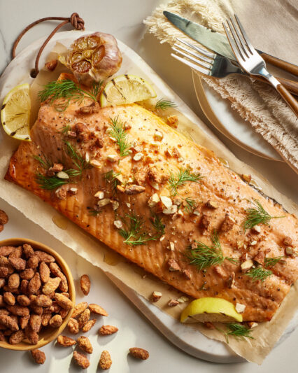 Roasted salmon fillet with fresh dill, chopped NaturSource almonds, lemon wedges, and roasted garlic, styled on parchment over a marble serving board.